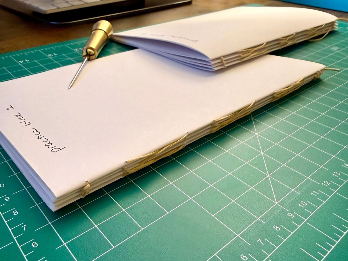 Two hand-stitched practice book blocks and a brass awl on a green cutting mat